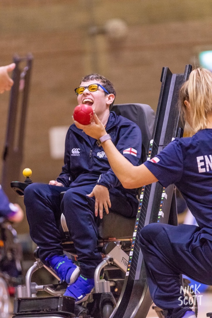 Beth Moulam, boccia player wearing England kit and coloured lens glasses, sat in power chair, red boccia ball being handed by assistant to referee