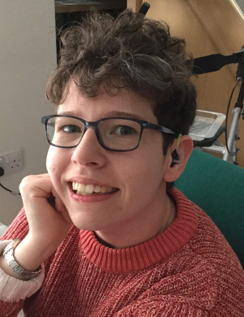 Beth Moulam woman with cerebral palsy, smiling, wears an orange jumper and blue glasses. 