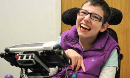 Beth Moulam woman with cerebral palsy. Wears a purple sleeveless jacket. Sat in electric power chair, in front a communication aid, AAC device is mounted on a stand at the front of the chair.