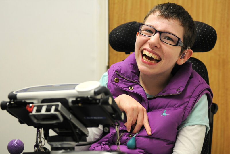 Beth Moulam woman with cerebral palsy. Wears a purple sleeveless jacket. Sat in electric power chair, in front a communication aid, AAC device is mounted on a stand at the front of the chair. Using a zip pull designed to help open and close a zip. Occupational therapy.