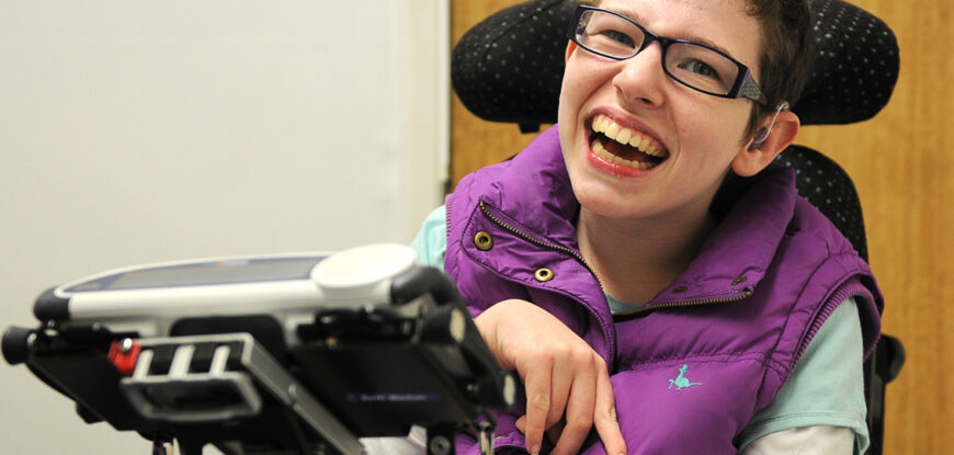 Beth Moulam woman with cerebral palsy. Wears a purple sleeveless jacket. Sat in electric power chair, in front a communication aid, AAC device is mounted on a stand at the front of the chair.