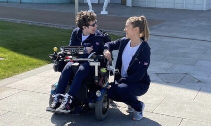 Beth Moulam sat in power chair outside using communication device, AAC. Talking with Christie, her boccia sports assistant who is crouching. Both wearing navy sports kit with Boccia UK logo.