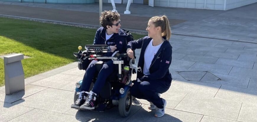 Beth Moulam sat in power chair outside using communication device, AAC. Talking with Christie, her boccia sports assistant who is crouching. Both wearing navy sports kit with Boccia UK logo.