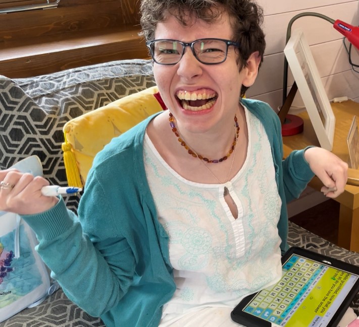 Beth Moulam woman in glasses wearing white blouse with turquoise embroidery with a turquoise cardigan. Holding a stylus in clenched hand to use with a communication aid on knee. Occupational therapy.