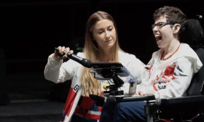 Beth Moulam in power chair using communication aid, sports assistant holding microphone to communication aid so Beth's voice can be heard at Leeds Arena