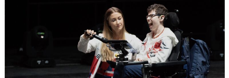 Beth Moulam in power chair using communication aid, sports assistant holding microphone to communication aid so Beth's voice can be heard at Leeds Arena