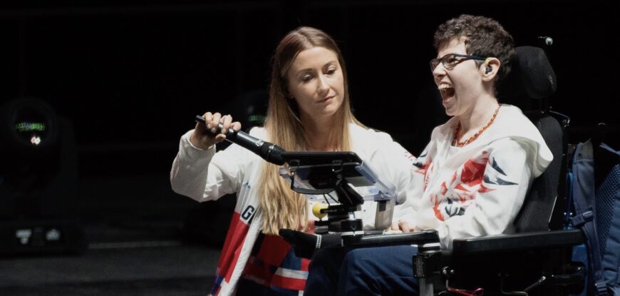 Beth Moulam in power chair using communication aid, sports assistant holding microphone to communication aid so Beth's voice can be heard at Leeds Arena