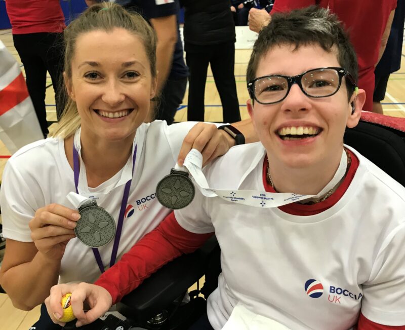 Beth Moulam and Christie Hutchings, cerebral palsy boccia athlete and assistant. Wearing white boccia UK kit and sporting silver medals. Beth has glasses and hearing aids.