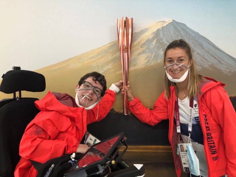 Beth Moulam in power chair and with communication aid, together with sports Assistant Christie Hutchings. Both women wearing GB rain jackets and holding the Paralympic Torch in the athlete village at the Tokyo 2020 Games 