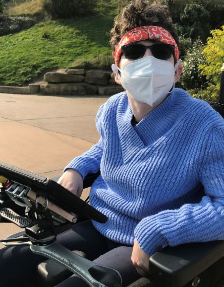 Beth Moulam, woman in wheelchair using a communication aid, wearing blue jumper, sunglasses and a mask held in position by a headband