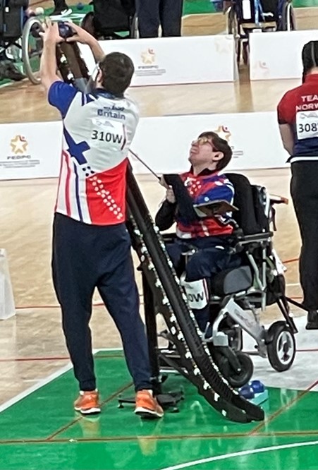 Beth Moulam BC3 boccia athlete wearing GB sports kit preparing to take shot. Ramp assistant with competitor number on back, under direction places the ball on the ramp ready for Beth to release.