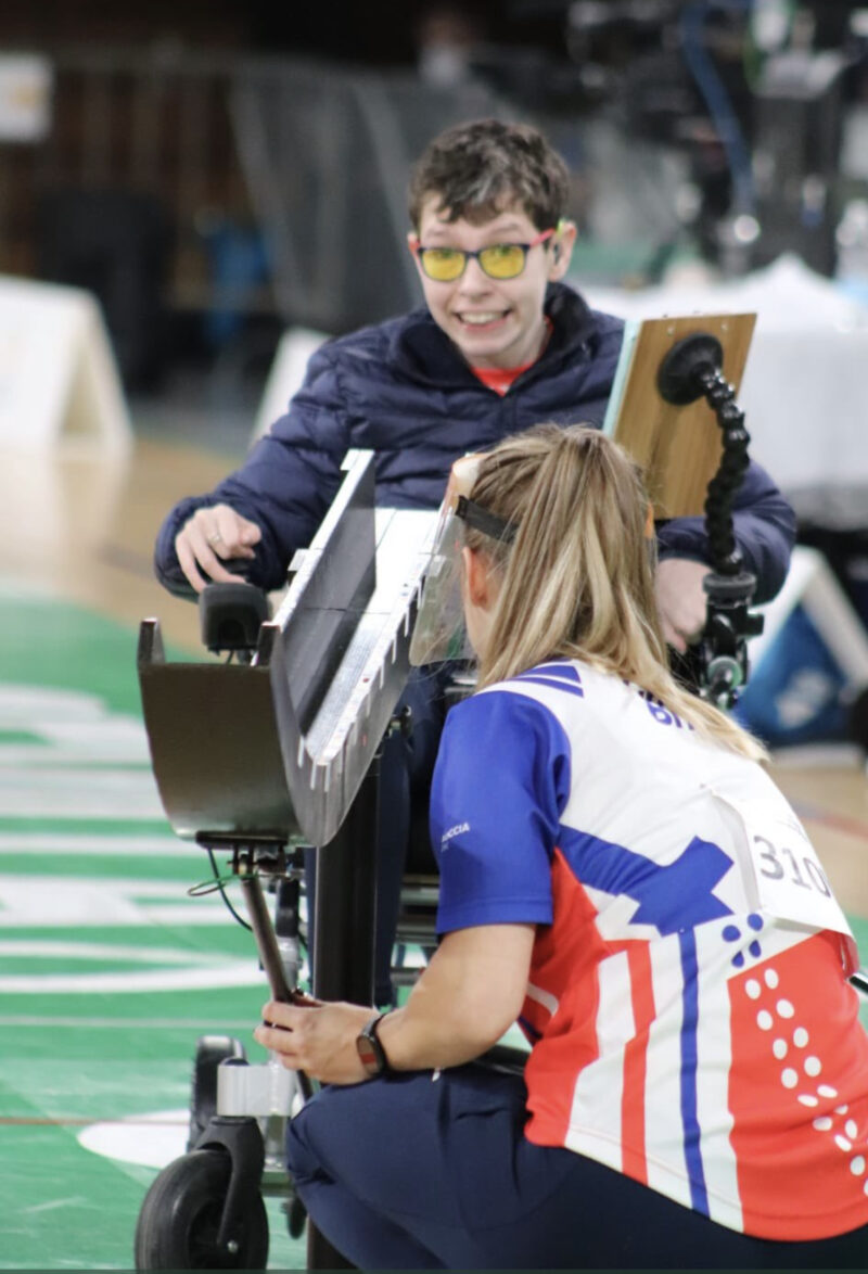 Beth Moulam BC3 boccia athlete wearing blue coat and yellow glasses lines up the ramp whilst Ramp Operator Christie Hutchings wearing multi-coloured shirt moves the ramp under direction