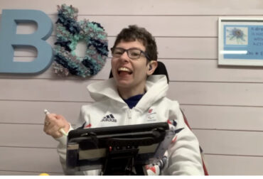 Beth Moulam holding stylus to direct input to communicate using AAC. Woman using AAC wearing glasses and sporting white sports top.