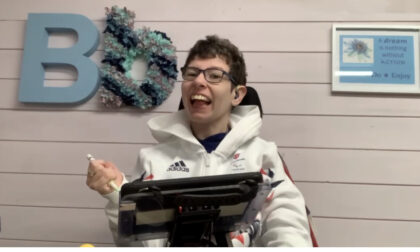 Beth Moulam holding stylus to direct input to communicate using AAC. Woman using AAC wearing glasses and sporting white sports top.