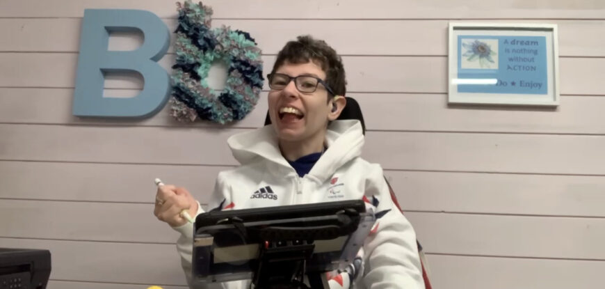 Beth Moulam holding stylus to direct input to communicate using AAC. Woman using AAC wearing glasses and sporting white sports top.