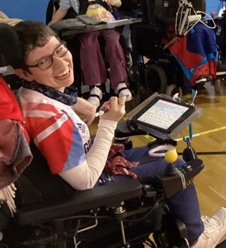 Beth Moulam wearing GB sports kit, sat in wheelchair with iPad and holding stylus. Working with group of teenagers.