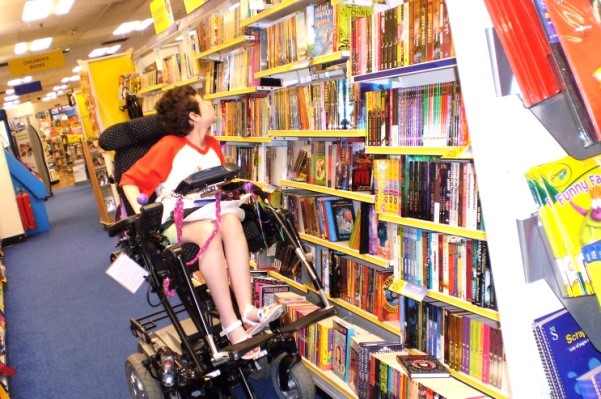 Beth Moulam, girl wearing red and white t-shirt and sandals, sat in power wheelchair, using riser on chair to look at high shelves in bookshop. International wheelchair day.