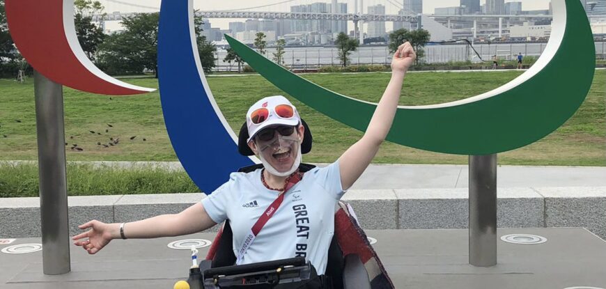 Beth Moulam, boccia athlete, in Tokyo 2020 Paralympic Games athlete village in front of the agitos logo. Wearing pale blue GB kit, hat and sunglasses on head. Reflections on stepping back from boccia