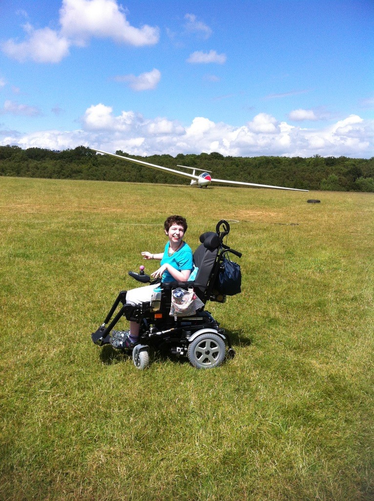 Beth Moulam, teenage girl, wearing turquoise t-shirt, sat in power wheelchair in field with glider behind, Blue sky with clouds. Celebrating international wheelchair day.