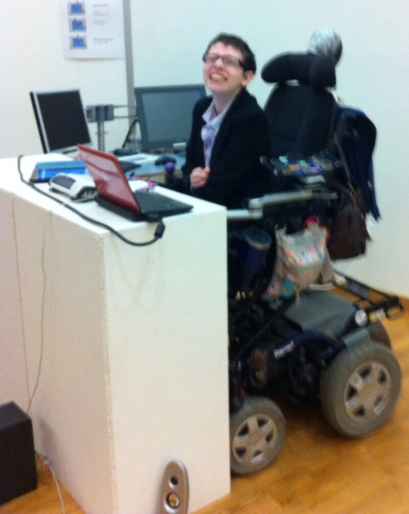 Beth Moulam, young woman wearing black and glasses, sat in power chair at a lectern. Chair raised to allow access to equipment on desk. Celebrating international wheelchair day.