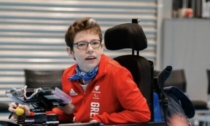 Beth Moulam, GB Boccia Paralympian 2020. Beth sits in power chair delivering a boccia training session. She has her communication aid, known as an AAC device, mounted on the front of her chair. Beth wears white trainers, navy bottoms and a red GB top. Beth is also wearing glasses.. The sports hall floor is blue and in the foreground is the hand of a player who has thrown a blue boccia ball onto the court. Beth has retired from boccia but life never stands still she is still involved in delivery of the sport.