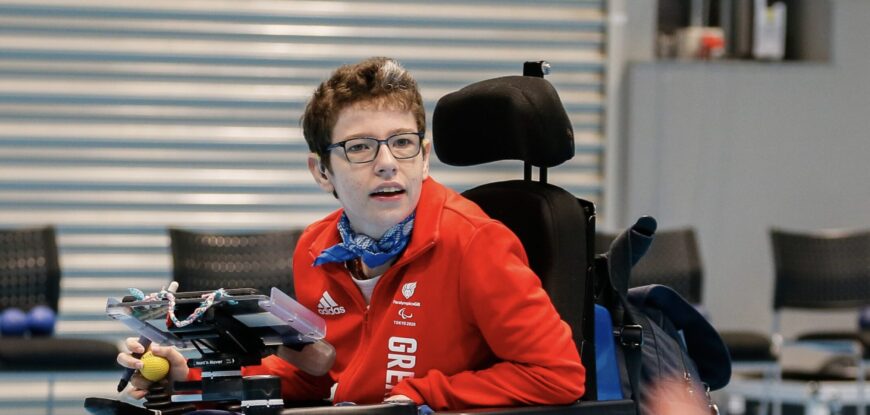 Beth Moulam, GB Boccia Paralympian 2020. Beth sits in power chair delivering a boccia training session. She has her communication aid, known as an AAC device, mounted on the front of her chair. Beth wears white trainers, navy bottoms and a red GB top. Beth is also wearing glasses.. The sports hall floor is blue and in the foreground is the hand of a player who has thrown a blue boccia ball onto the court. Beth has retired from boccia but life never stands still she is still involved in delivery of the sport.