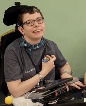 Beth Moulam, AAC role model for 1 Voice Charity. Beth wears a grey t-shirt with role model embroidered on the top. She is also wearing a scarf and glasses and sat in a power wheelchair. Beth holds a stylus used to access an electronic communication aid.