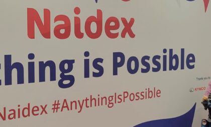 Beth Moulam at Naidex 2022. Beth sits in wheelchair in front of Naidex poster anything is possible. Ableism and communication impairment.