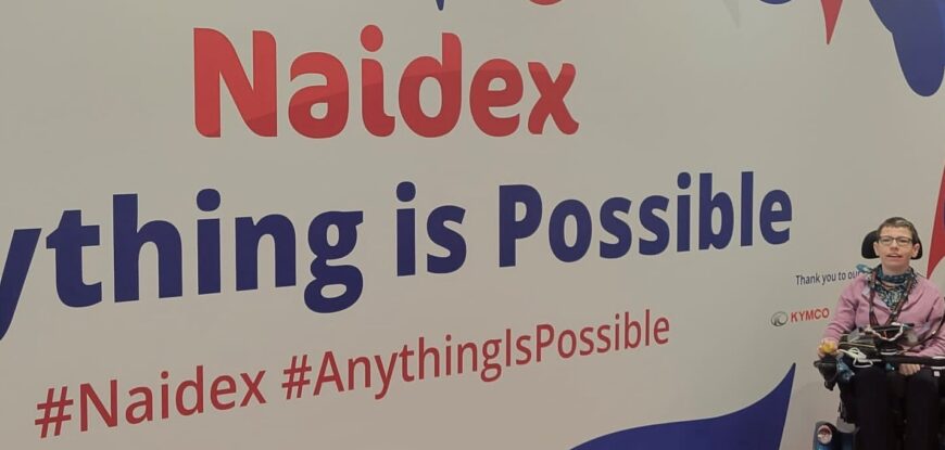 Beth Moulam at Naidex 2022. Beth sits in wheelchair in front of Naidex poster anything is possible. Ableism and communication impairment.