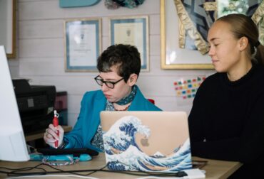 Beth and communication partner sat at desk decision making. Beth using a stylus with an ipad to communicate. AAC Ally