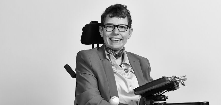 Beth Moulam Black and white photo sat in power chair with communication aid AAC device. Shaw Trust Disability Power 100 2023 Community Advocate