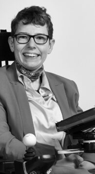 Beth Moulam Black and white photo sat in power chair with communication aid AAC device. Shaw Trust Disability Power 100 2023 Community Advocate