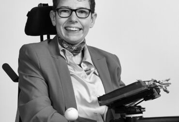 Beth Moulam Black and white photo sat in power chair with communication aid AAC device. Shaw Trust Disability Power 100 2023 Community Advocate